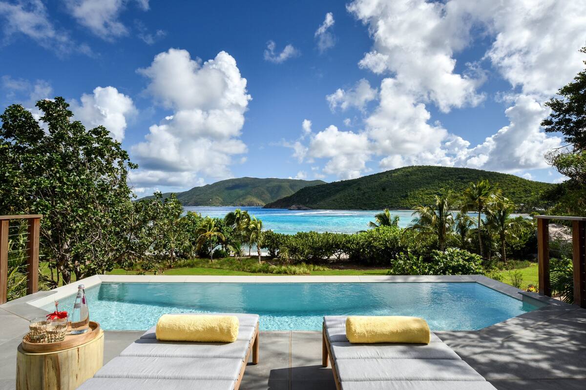 Une piscine bordée de deux chaises longues, entourée d’une végétation luxuriante et offrant une vue sur l’océan et les montagnes.