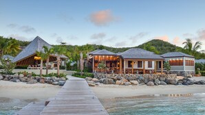 Un resort con una terraza de madera que da a una playa, con varias cabañas sobre el agua, techos de paja y paredes de piedra.
