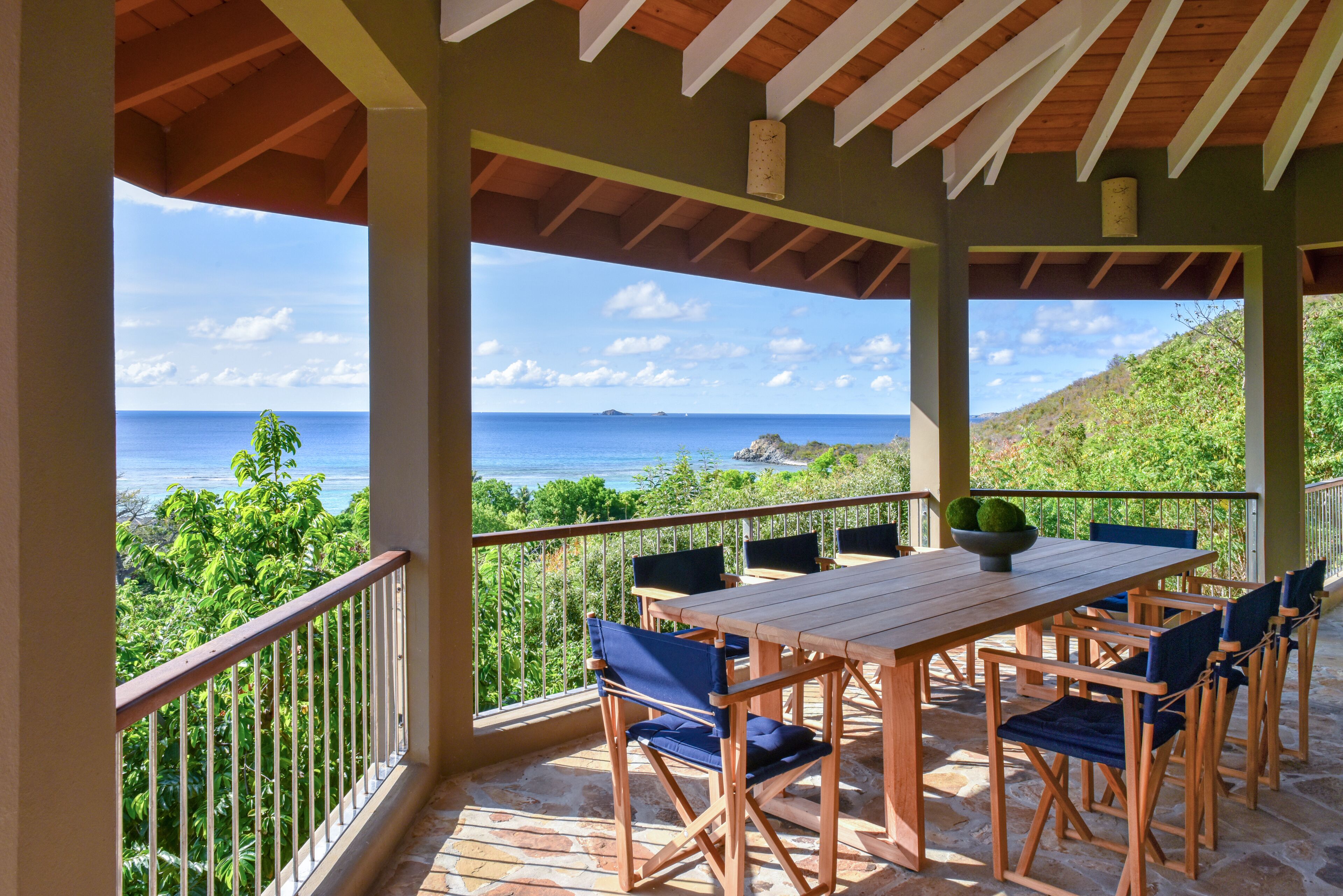 A covered outdoor seating area with a table and chairs, overlooking a scenic view of the ocean and greenery.