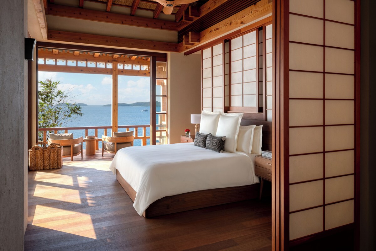 A bedroom with a large bed, wooden flooring, a view of the sea, and a traditional Japanese-style room divider.