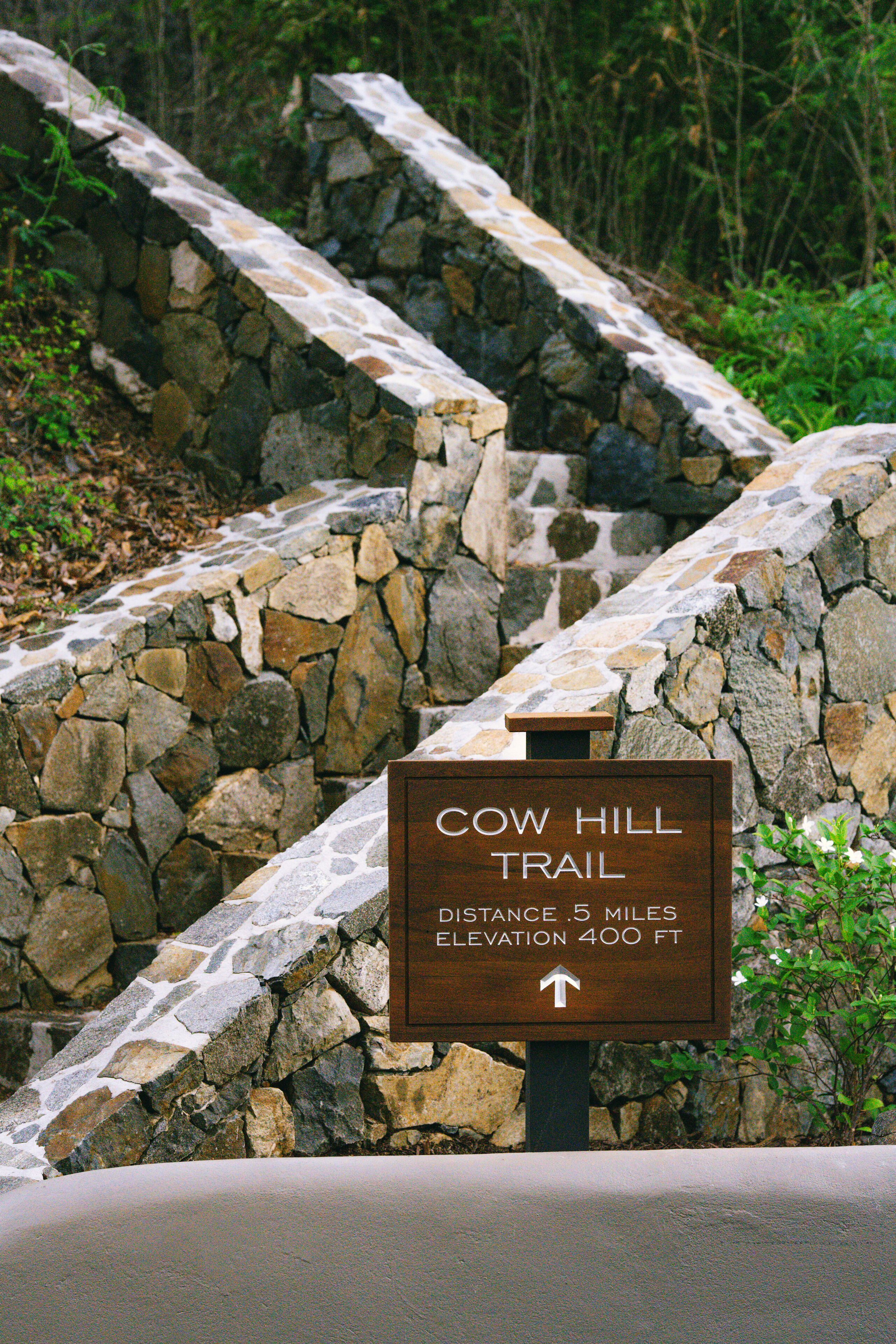 Una escalera de piedra que conduce a un cartel del Sendero Cow Hill, que indica una distancia de 8 kilómetros y una ganancia de elevación de 120 metros.