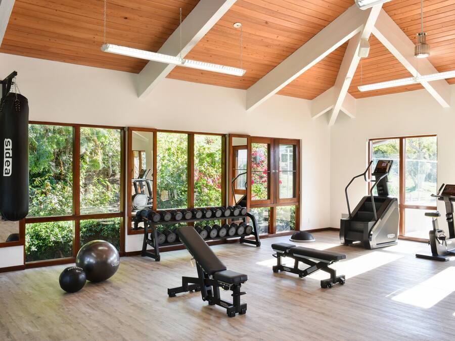 A spacious gym with wooden floors, large windows, and various exercise equipment including dumbbells, a bench, and treadmills.