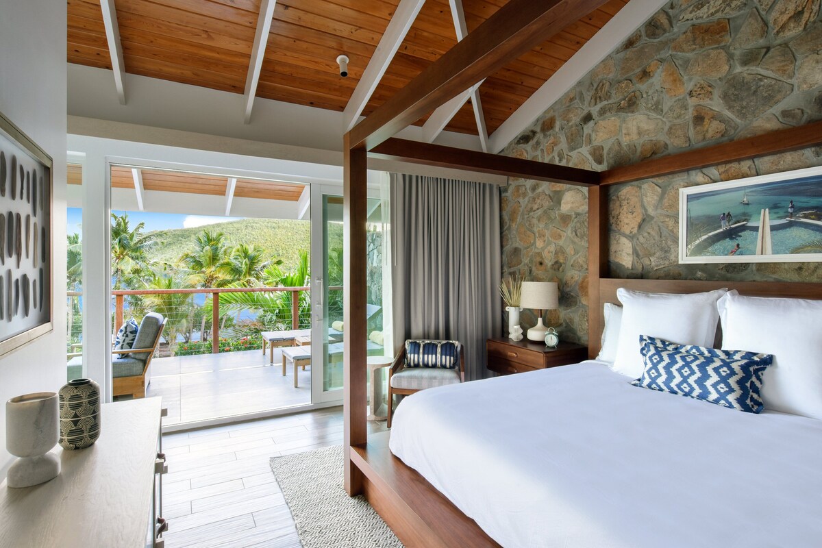 A bedroom with a large bed, a wooden ceiling, stone walls, a balcony with a view of greenery, and a painting on the wall.