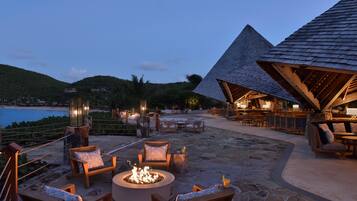 Un patio exterior con suelo de piedra, muebles de madera y un pozo de fuego.