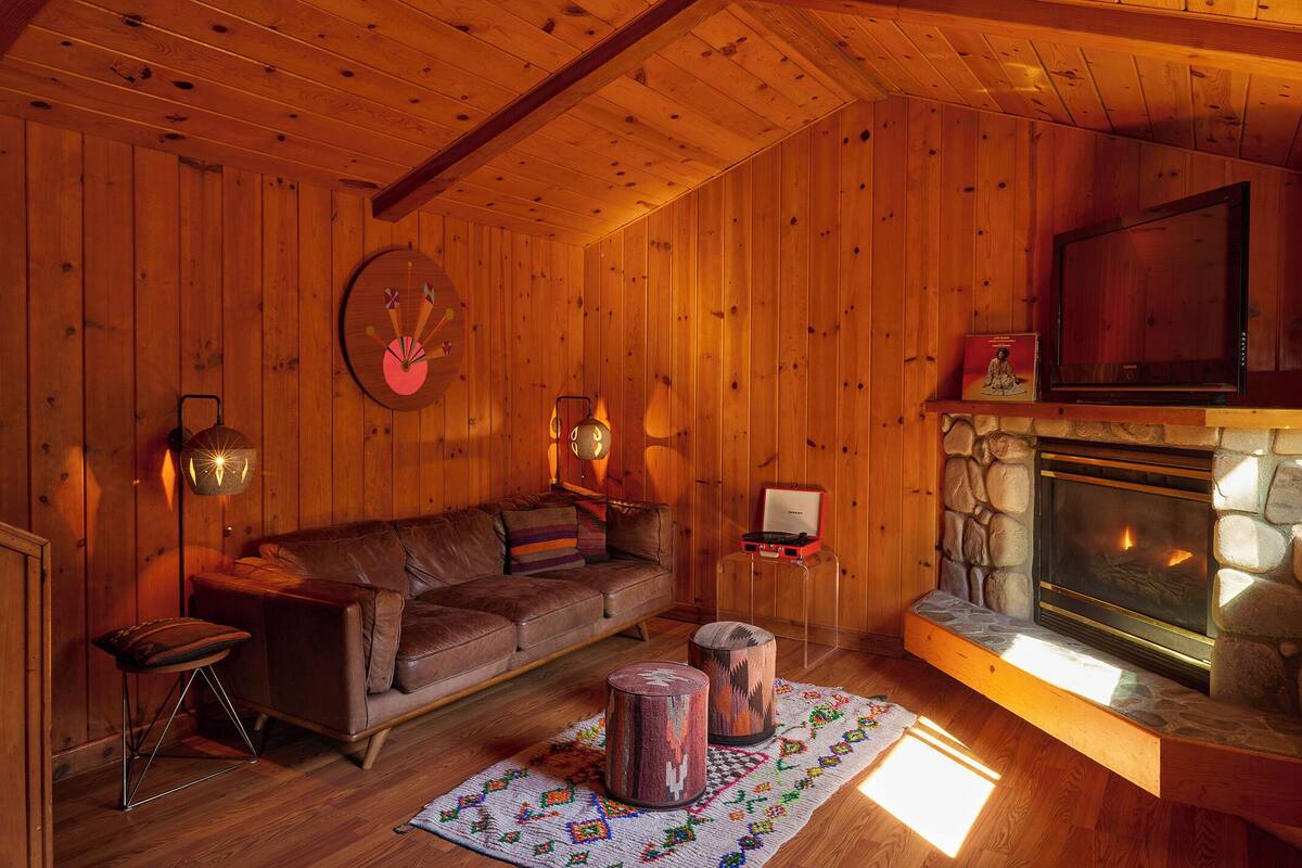 A cozy cabin interior with a stone fireplace, wooden walls, a leather sofa, and a television.