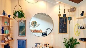 A room with white walls, wooden ceiling beams, shelves with various items, a round mirror, and a small bench with orange towels.