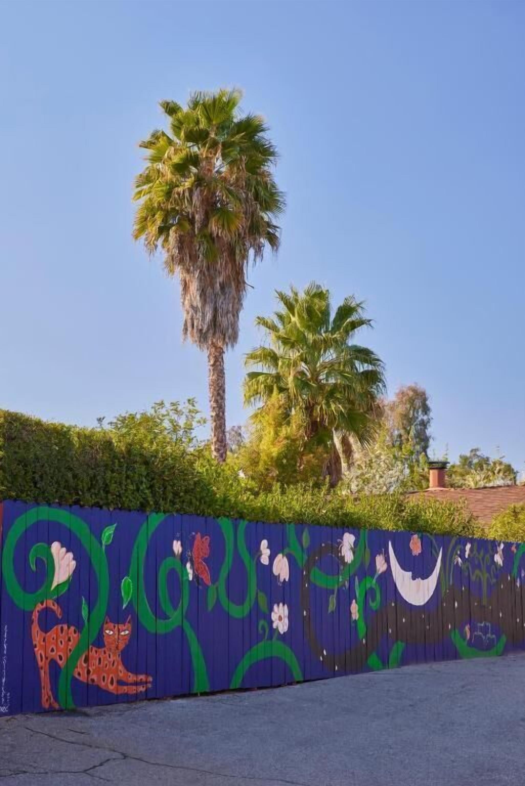 A fence with colorful floral and animal designs, palm trees, and a clear blue sky.