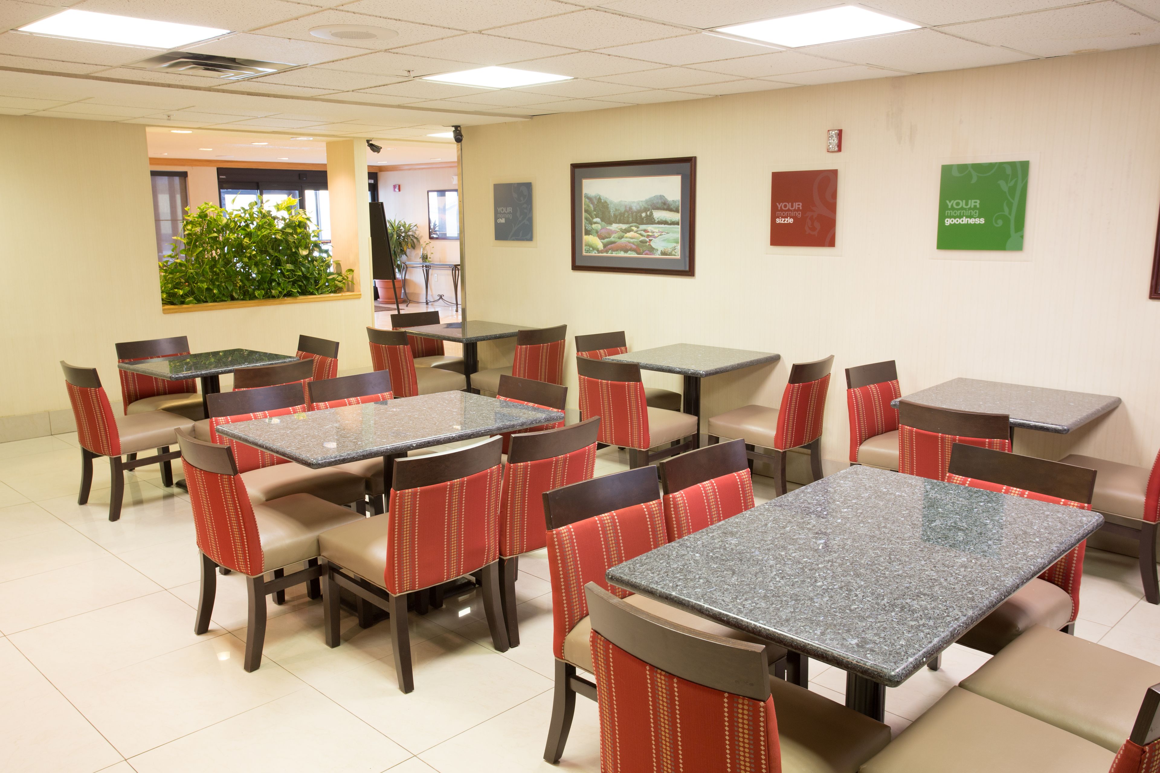 A hotel dining area with multiple tables and chairs, a potted plant, and framed pictures on the walls.
