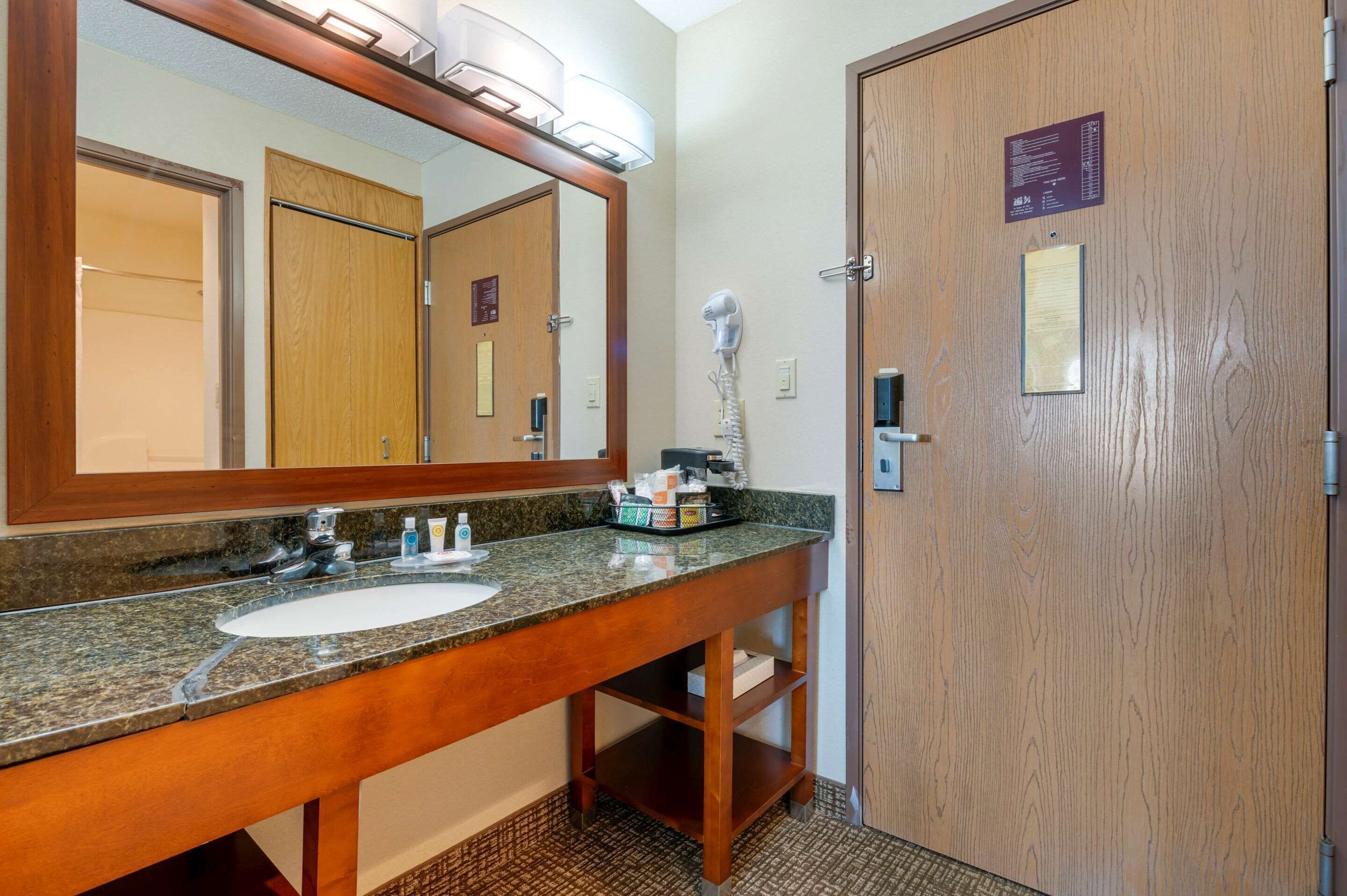 A bathroom with a granite countertop sink, a large mirror, and a hairdryer on the wall. There are wooden doors and a small shelf under the sink.