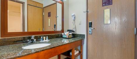 A bathroom with a granite countertop sink, a large mirror, and a hairdryer on the wall. There are wooden doors and a small shelf under the sink.
