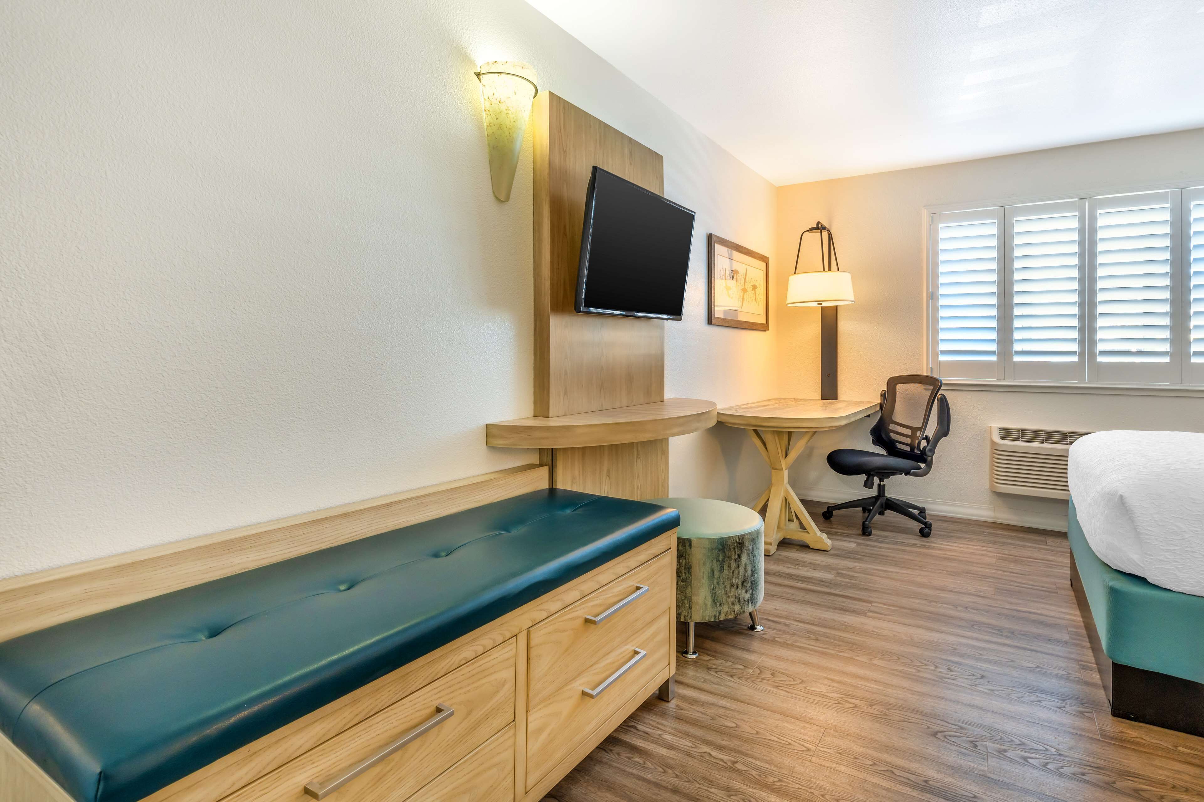 A hotel room with a wooden bench, a desk, a chair, a bed, a TV, and a window with blinds.