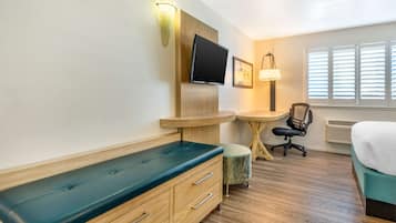 A hotel room with a wooden bench, a desk, a chair, a bed, a TV, and a window with blinds.