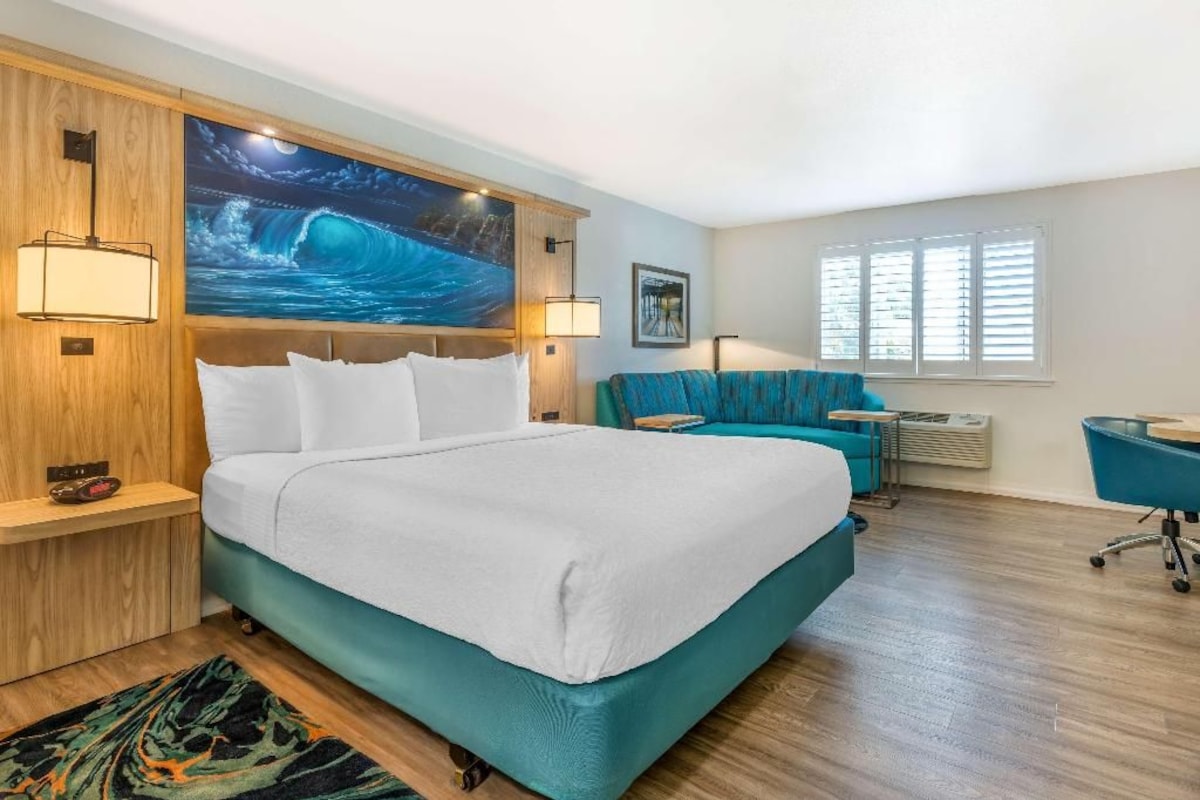 A hotel room with a large bed, a desk, a chair, and a painting on the wall.