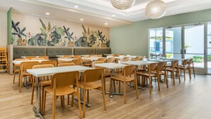 A spacious dining area with wooden tables and chairs, a mural of tropical plants on the wall, and large windows offering a view of the outdoors. - Best Western Plus Capitola By-the-sea Inn & Suites (Santa Cruz)