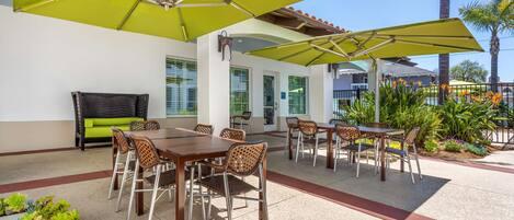 An outdoor patio with tables and chairs under green umbrellas, surrounded by plants and palm trees.