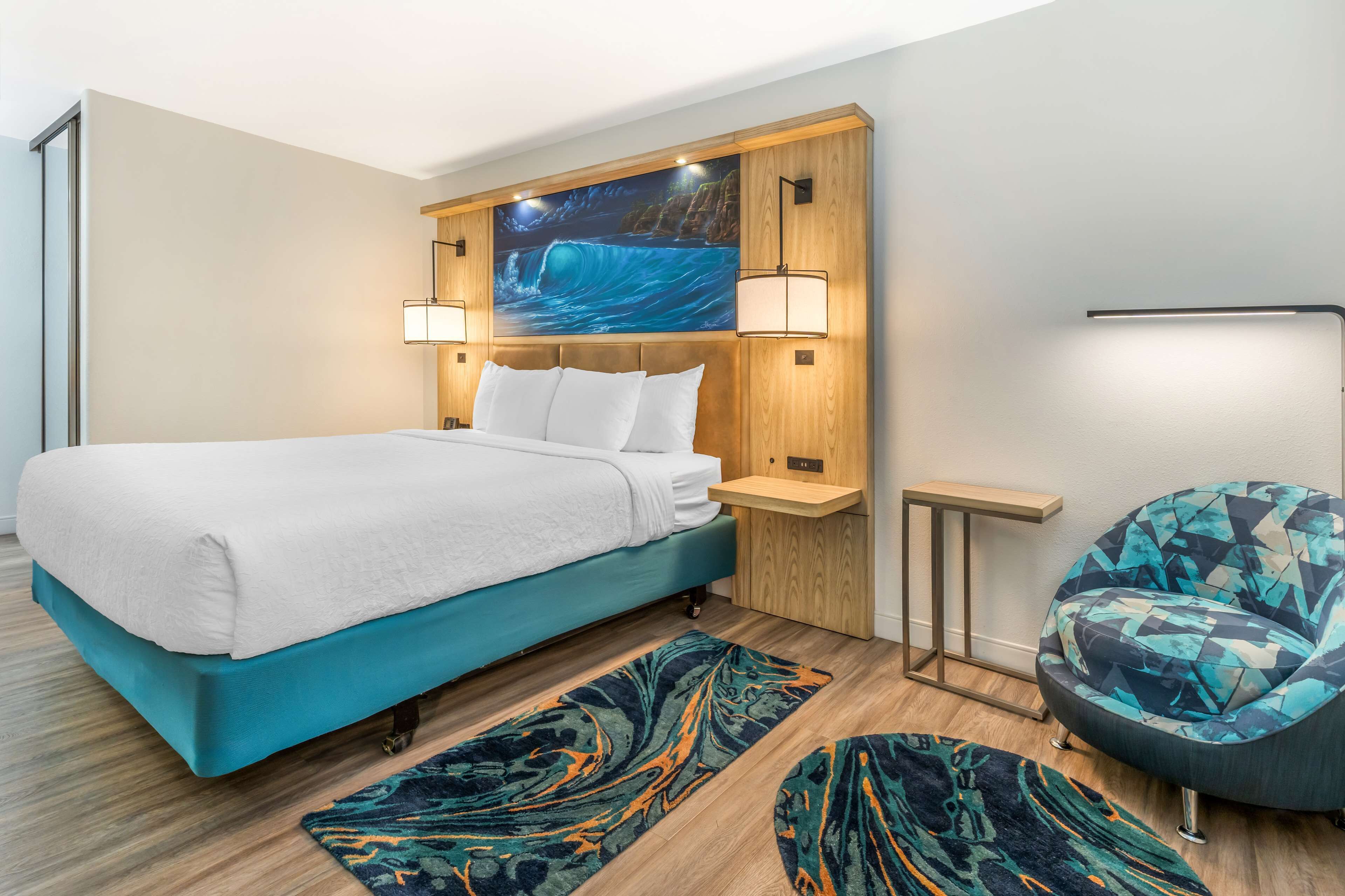 A modern hotel room with a large bed, a wooden headboard, a wall-mounted TV, a side table, a chair, and a patterned rug.