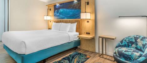 Una habitación de hotel moderna con una cama grande, cabecera de madera, televisor montado en la pared, mesita auxiliar, una silla y un tapete estampado.