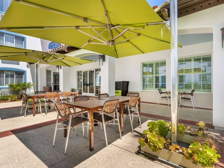 Um pátio externo com mesa e cadeiras de madeira, guarda-sóis verdes e plantas em vasos.