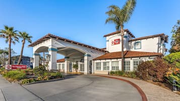 Welcome to our hotel, set amidst lush landscaping and palm trees, with a traditional facade and a wide, smooth entrance pathway.