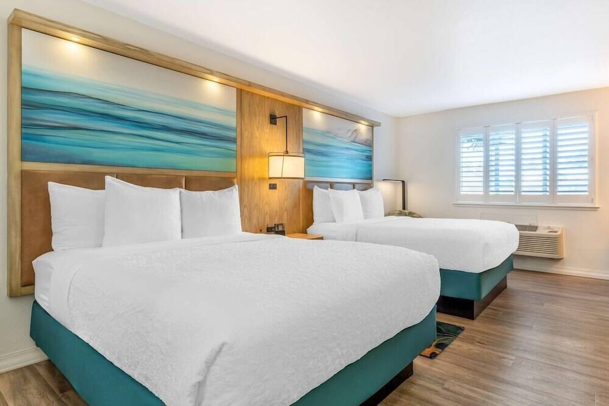 A hotel room with two beds, a wooden headboard, a large abstract painting, a window with blinds, and a wall-mounted air conditioning unit.