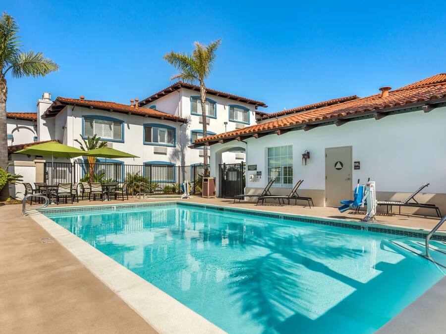 A swimming pool with lounge chairs and umbrellas, surrounded by buildings with tiled roofs and palm trees.