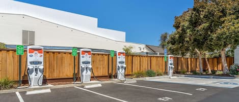 An EV charging station area with three charging points, a wooden fence, and a building in the background.
