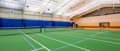 An indoor tennis court with a net, blue curtains, and a high ceiling with hanging lights.