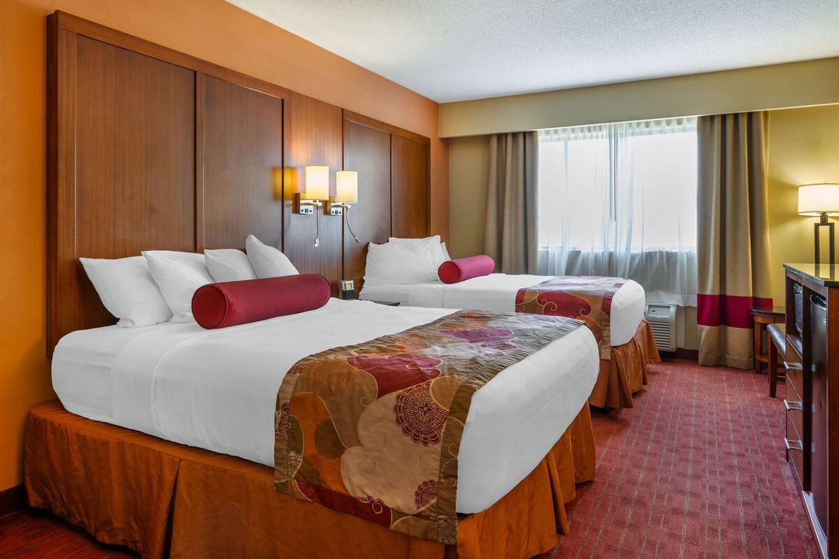 Habitación de hotel con dos camas, cabecera de madera, dos lámparas de noche, un escritorio y una ventana con cortinas.