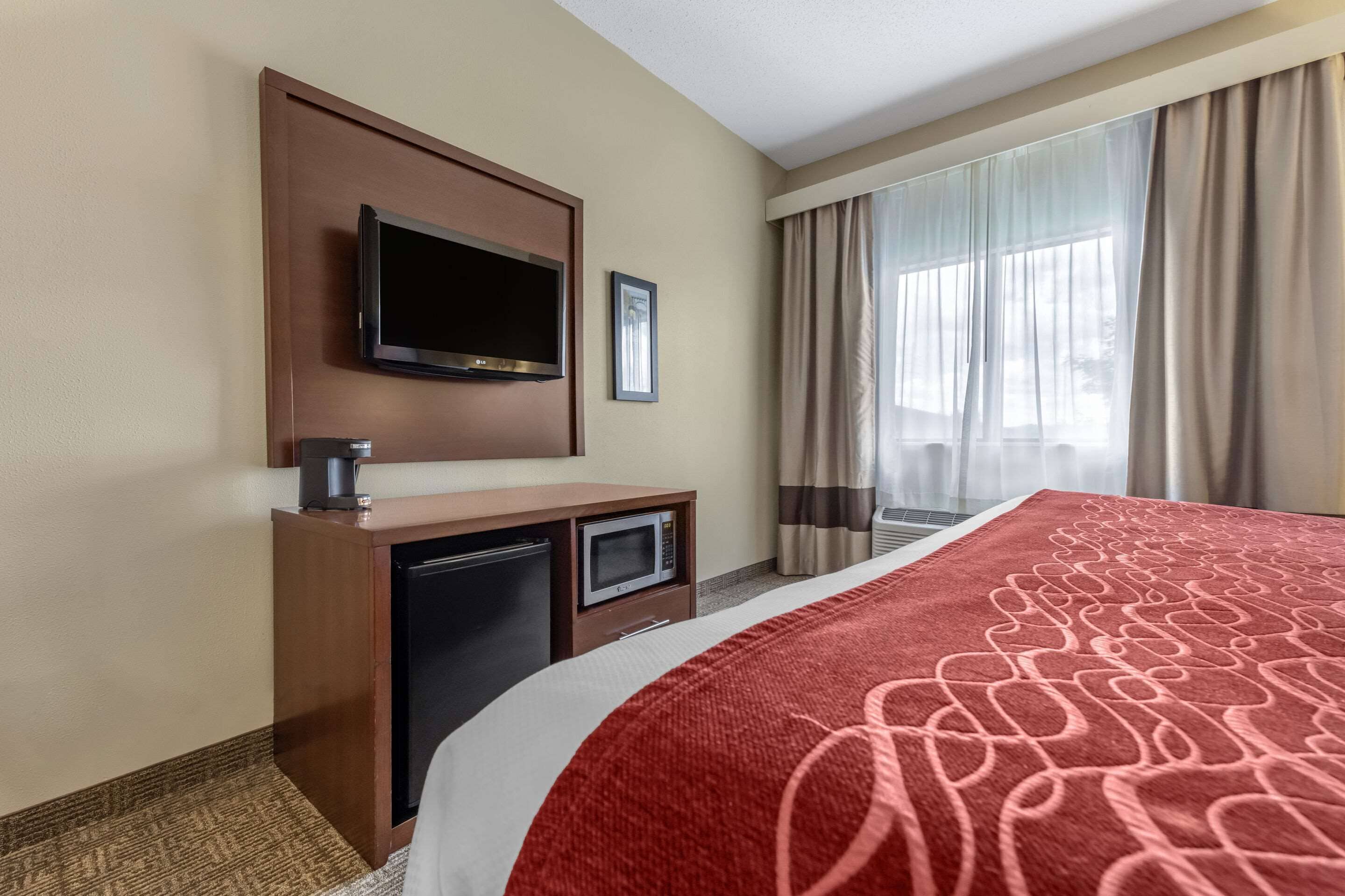 A hotel room with a bed, a television mounted on the wall, a small desk with a microwave, and a refrigerator.