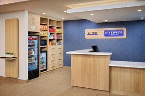 A hotel reception area with a 'Welcome' sign, a 'City Express by Marriott' sign, a refrigerator stocked with beverages, and a snack shelf.