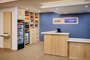 Zona de recepción de un hotel con un cartel de 'Welcome', un letrero de 'City Express by Marriott', un refrigerador con bebidas y una estantería con snacks.