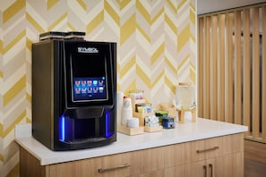 A coffee machine with a digital interface on a countertop, surrounded by coffee cups and containers. - City Express by Marriott Port Hope Cobourg (Port Hope)