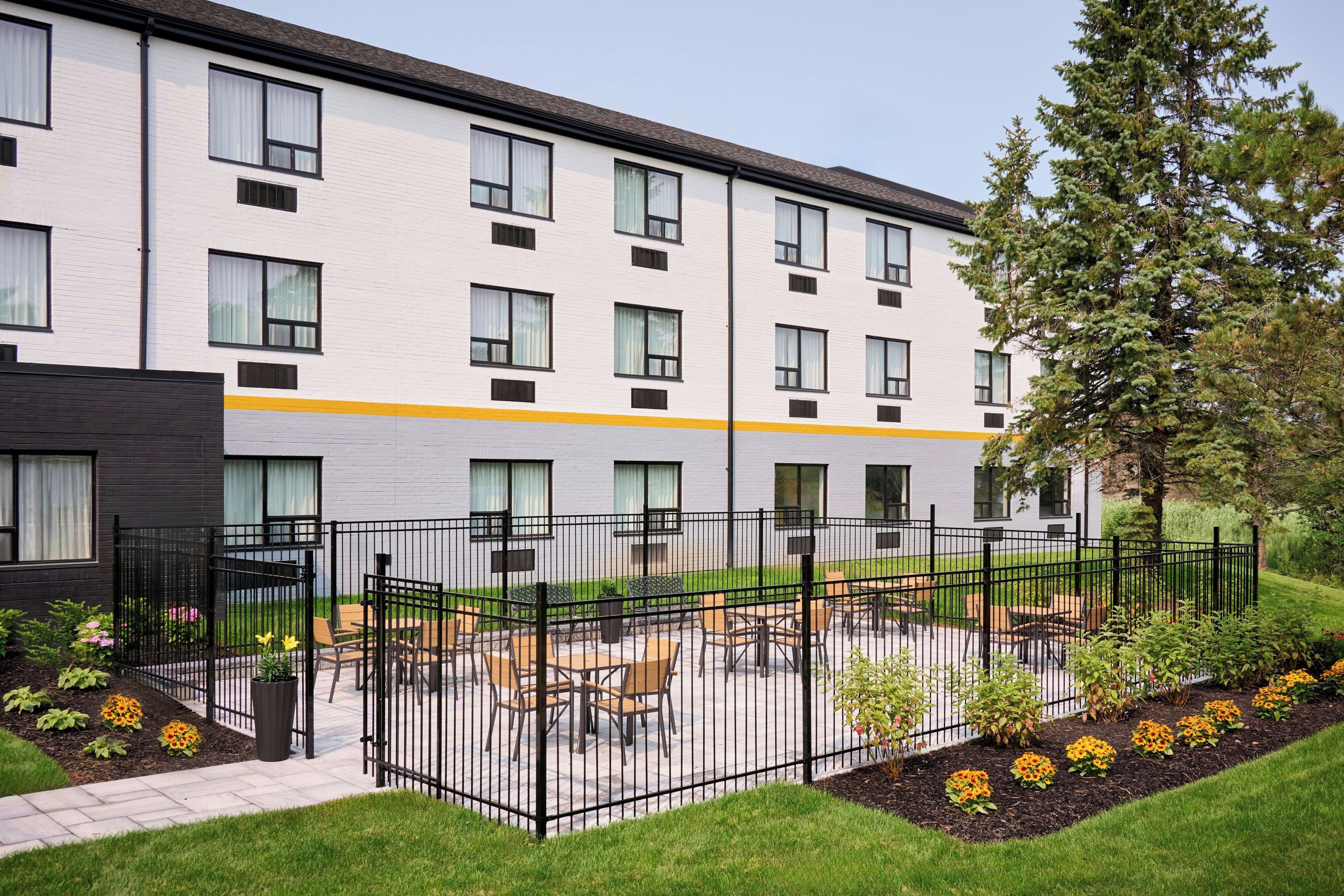 A multi-story building with a flat roof, a gated outdoor area with tables and chairs, and a well-maintained lawn with flower beds.