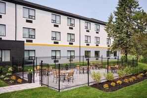 A multi-story building with a flat roof, a gated outdoor area with tables and chairs, and a well-maintained lawn with flower beds.