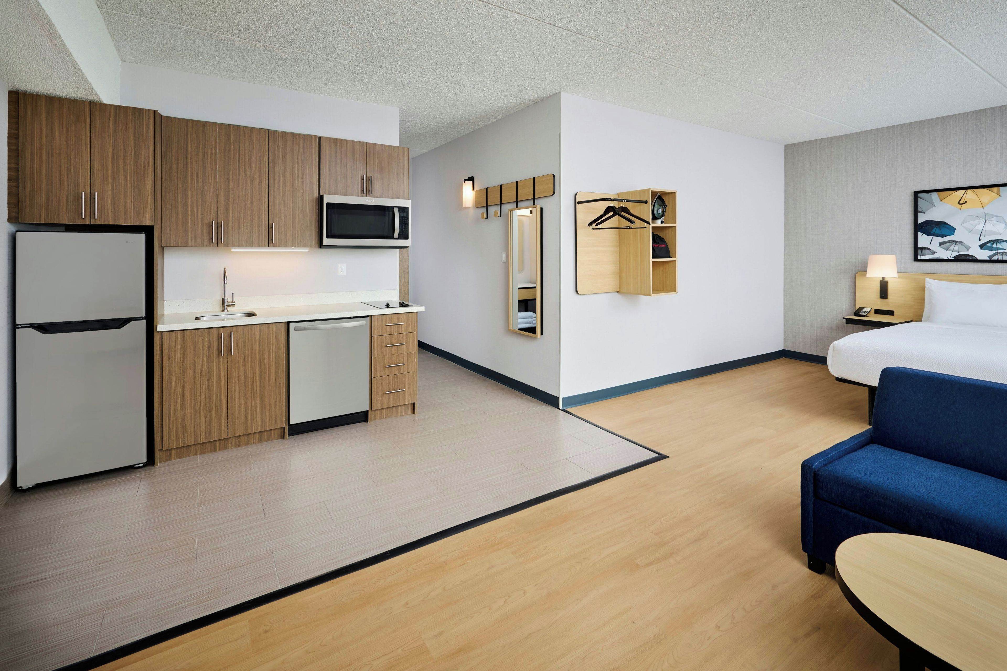 A compact hotel room with a kitchenette, a bed, a seating area, and a mirror.