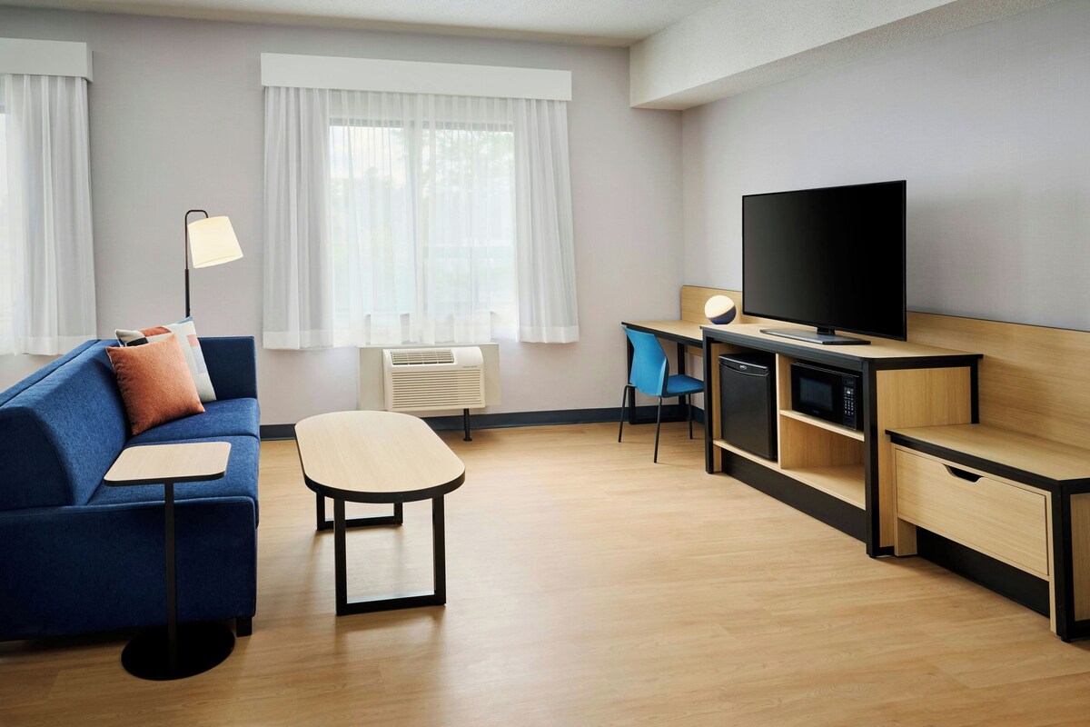 Une chambre d’hôtel moderne avec un canapé bleu, une petite table, un téléviseur, un bureau avec une chaise et un four à micro-ondes.