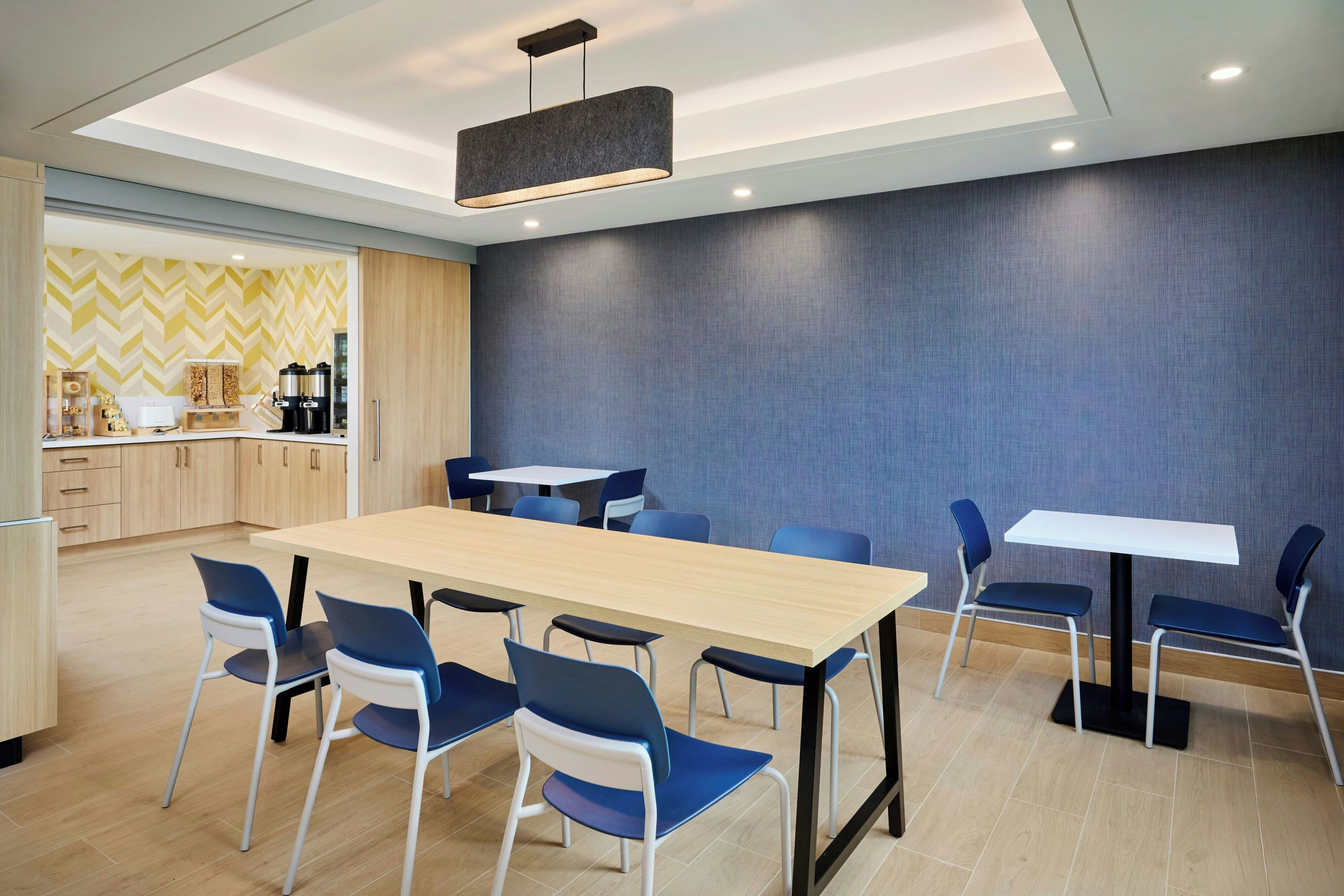 A modern dining area with a long table and blue chairs, a kitchen area with wooden cabinets, and a patterned wall.