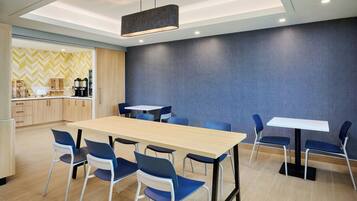 A modern dining area with a long table and blue chairs, a kitchen area with wooden cabinets, and a patterned wall.
