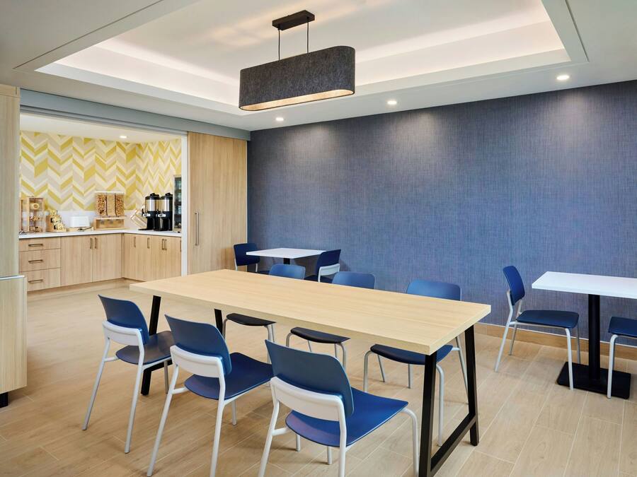 A modern dining area with a long table and blue chairs, a kitchen area with wooden cabinets, and a patterned wall.