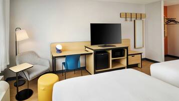 Habitación de hotel con una cama, un escritorio con televisor, una silla, una lámpara y un pequeño mueble para guardar objetos.