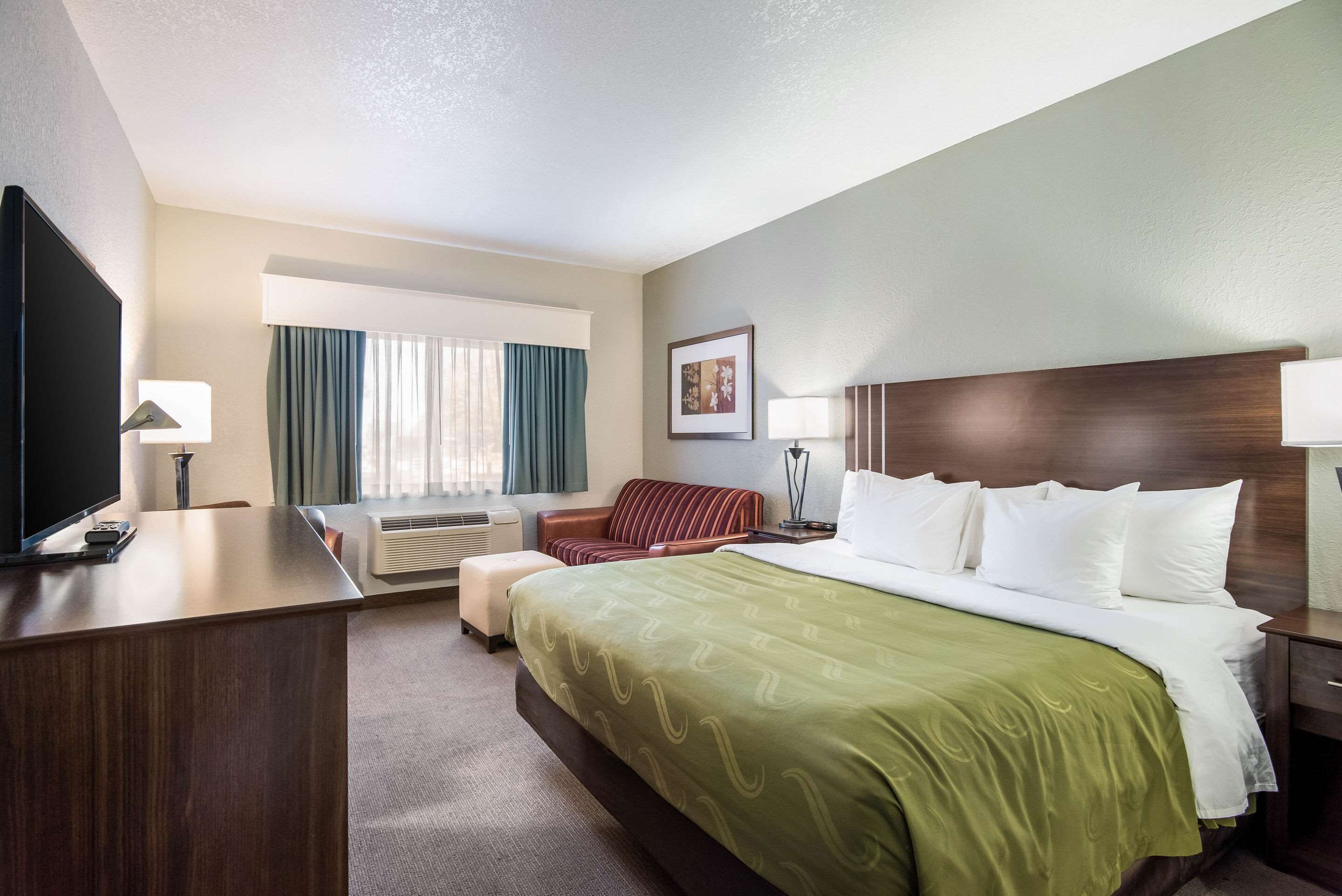 A hotel room with a large bed, a desk, a TV, a sofa, and a window with curtains.