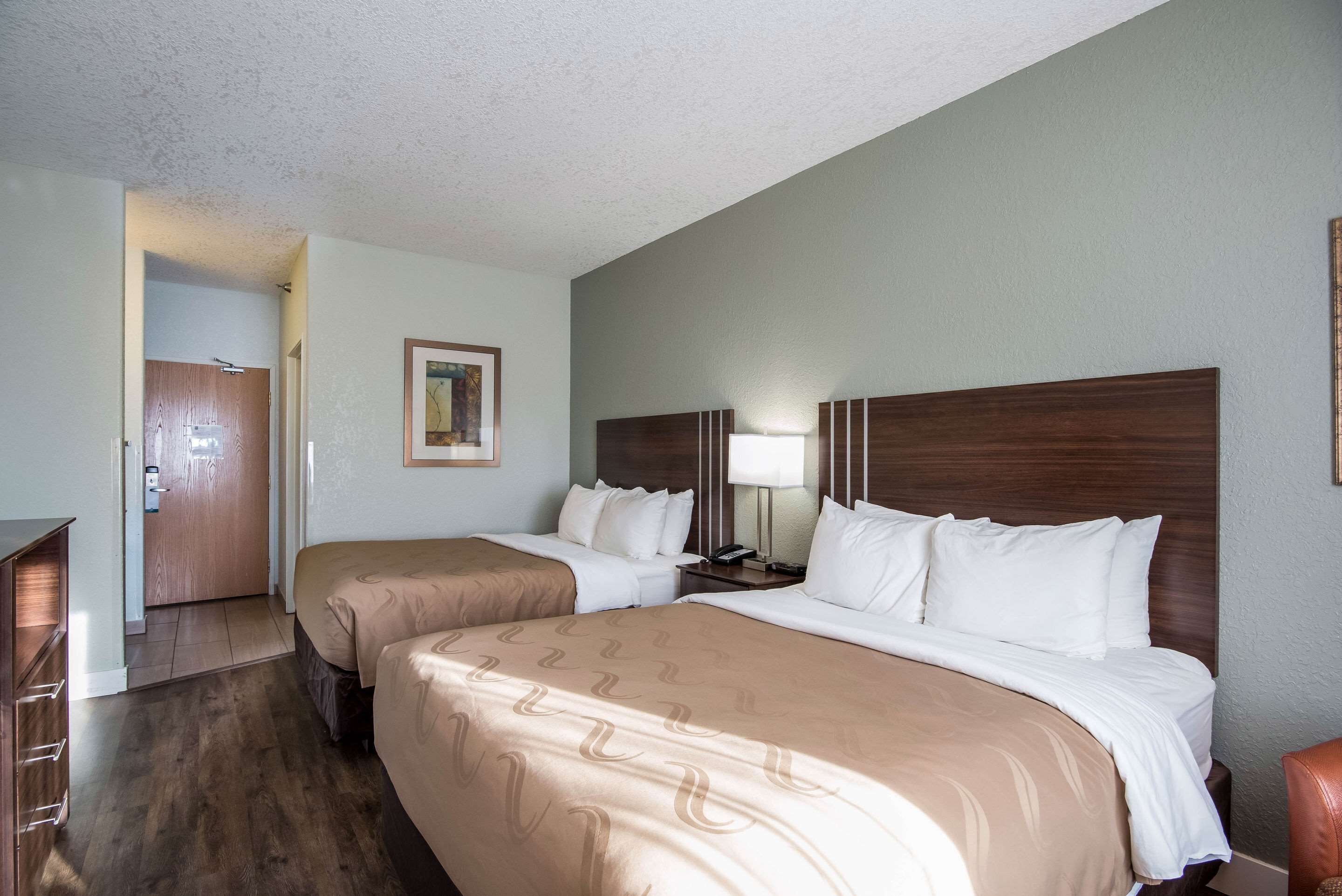 A hotel room with two beds, a wooden headboard, a nightstand with a lamp, a framed picture on the wall, and a wooden door leading to another room.