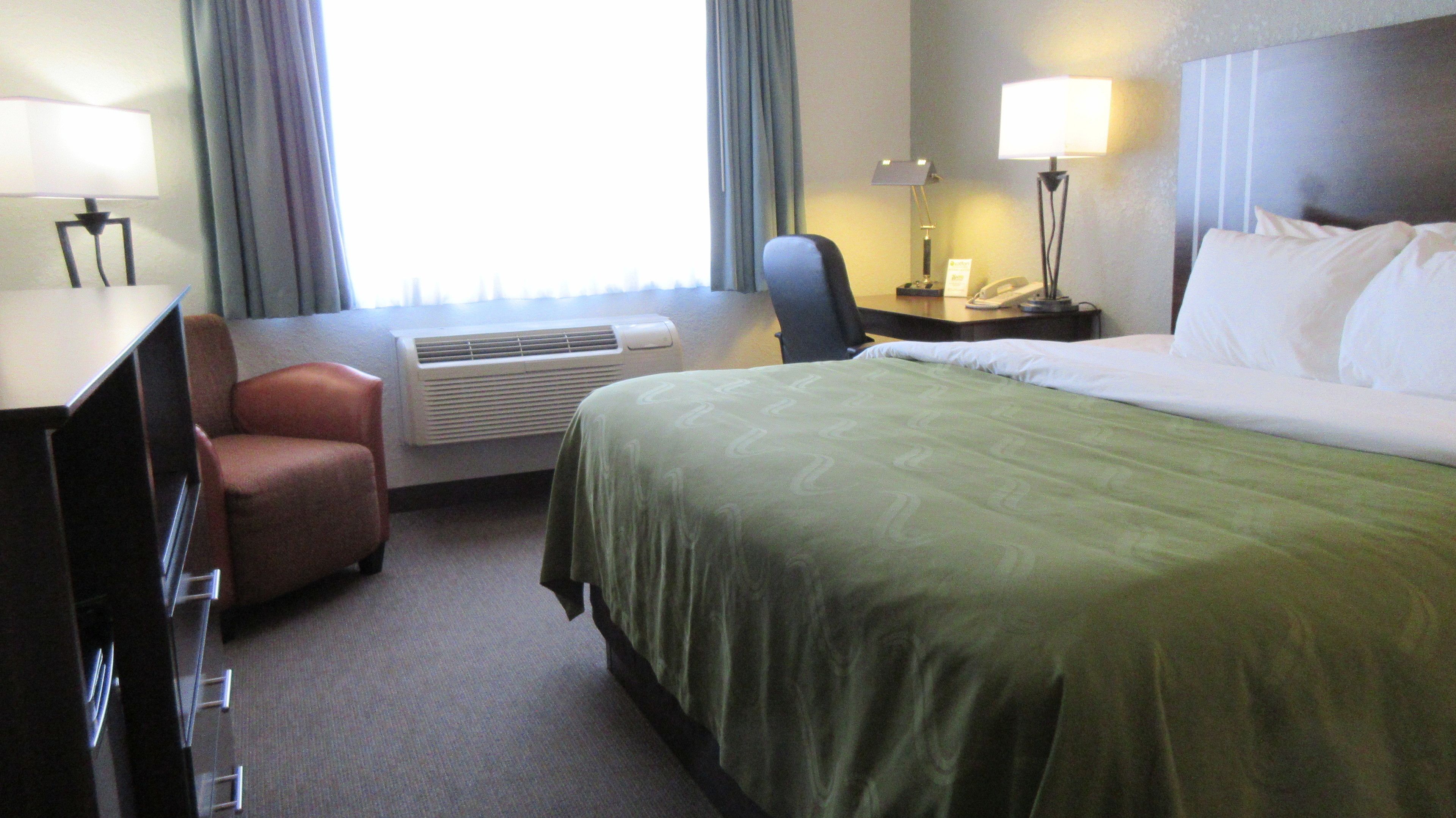 A hotel room with a bed, a desk, a chair, an air conditioning unit, and a telephone.