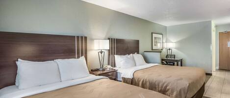 A hotel room with two beds, a nightstand with a lamp, a small table with a lamp, and a door.
