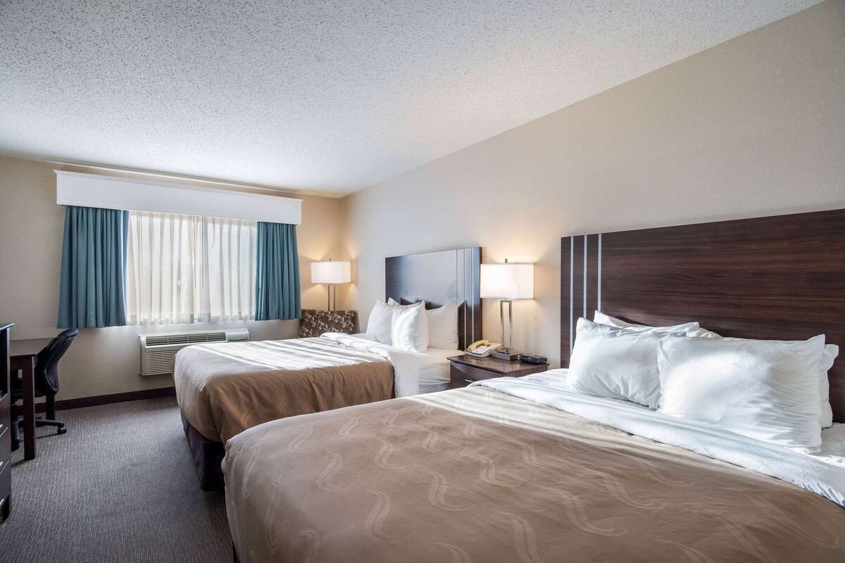A hotel room with two beds, a desk, a chair, and a large window with curtains.