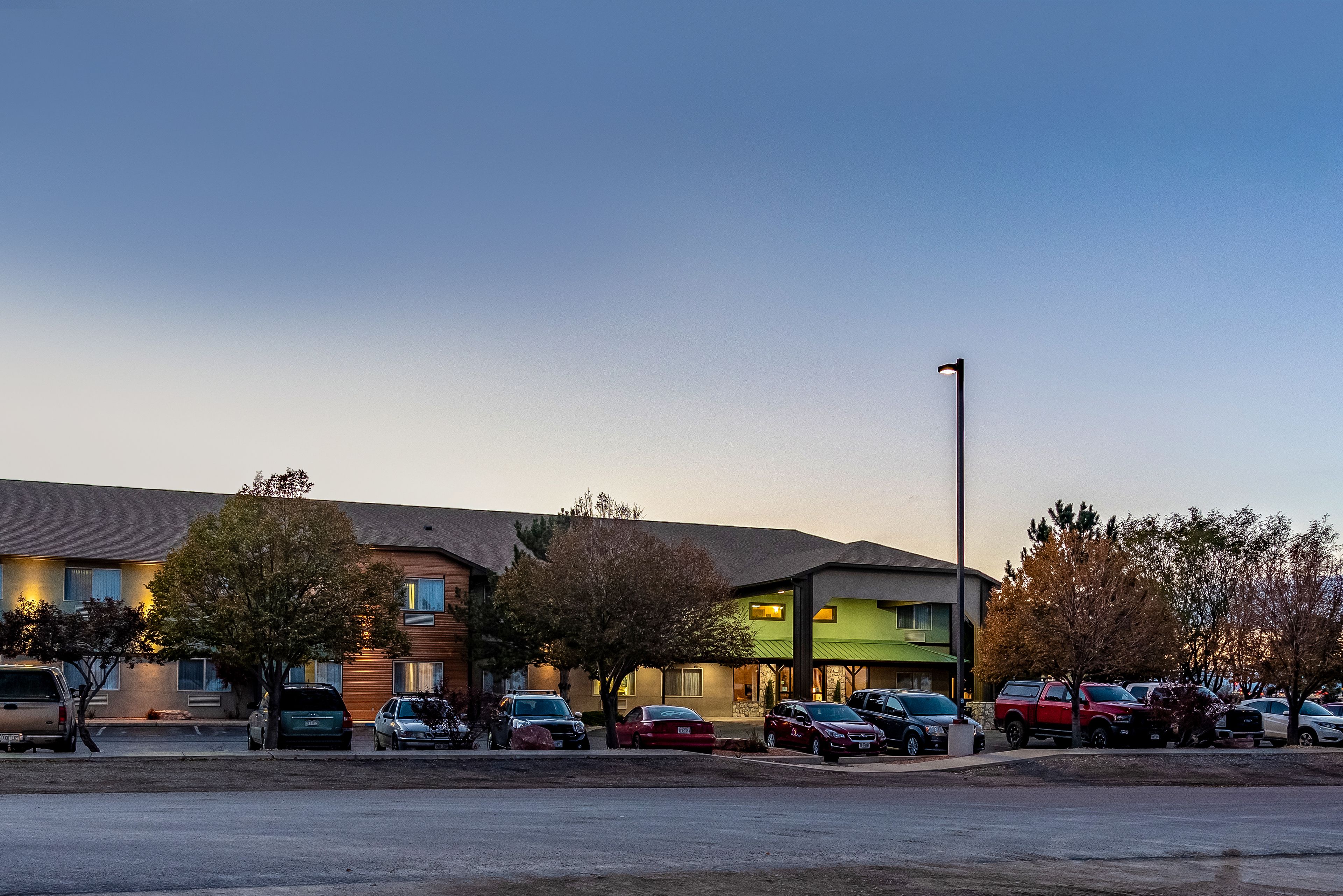 A parking lot with several cars and a building with multiple floors and a green-lit entrance.