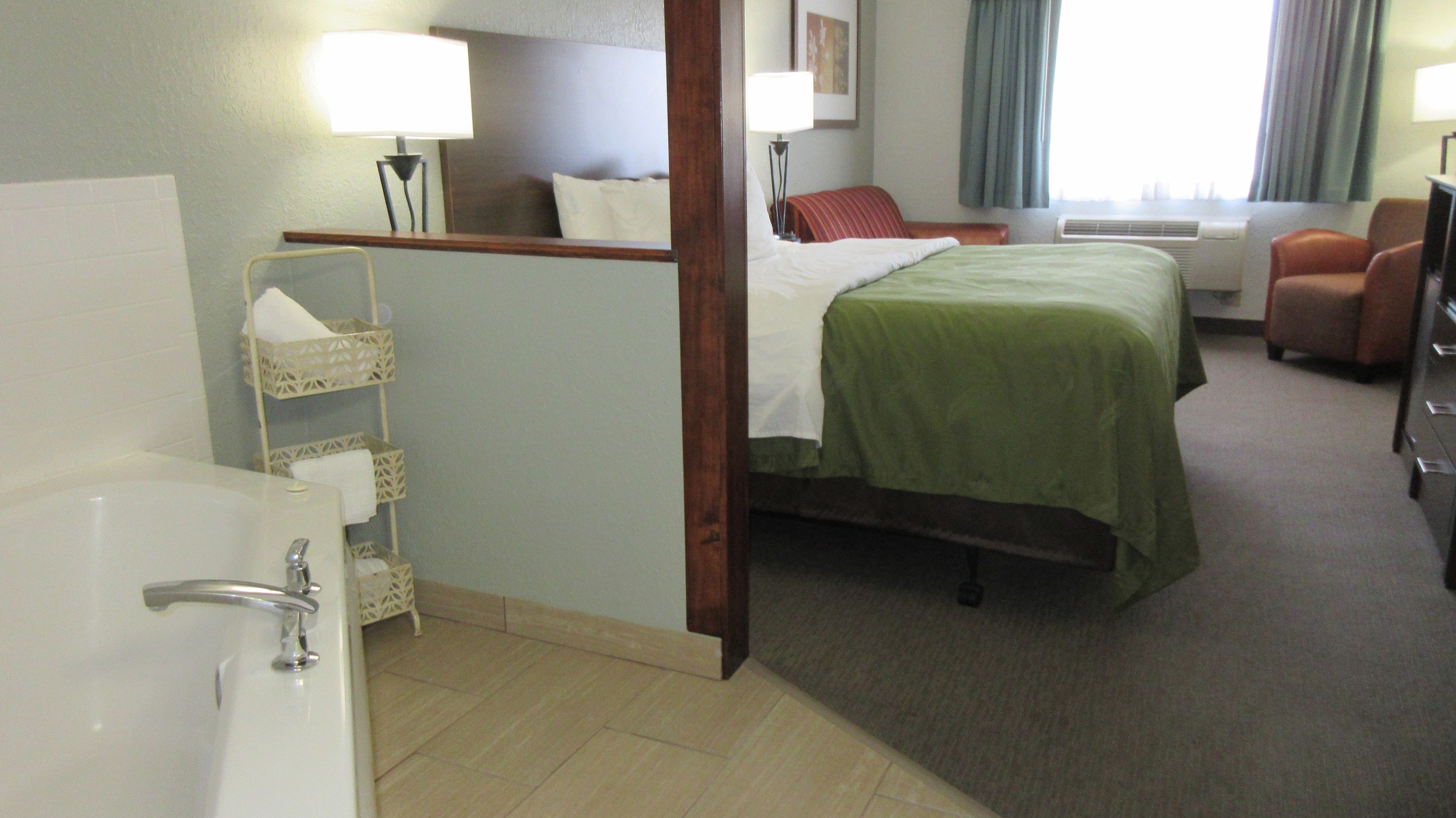 Ein Hotelzimmer mit einem Bett, einem Sessel, einem Schreibtisch, einer Lampe, einem Badezimmer mit einer Badewanne und einem Fenster mit Vorhängen.