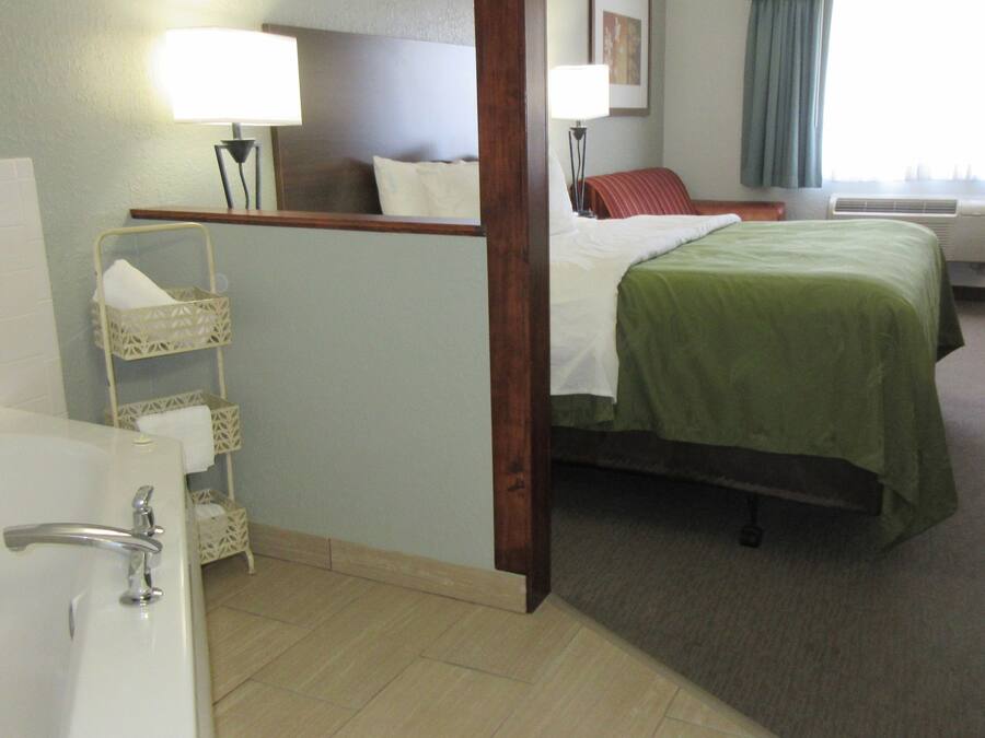 Ein Hotelzimmer mit einem Bett, einem Sessel, einem Schreibtisch, einer Lampe, einem Badezimmer mit einer Badewanne und einem Fenster mit Vorhängen.