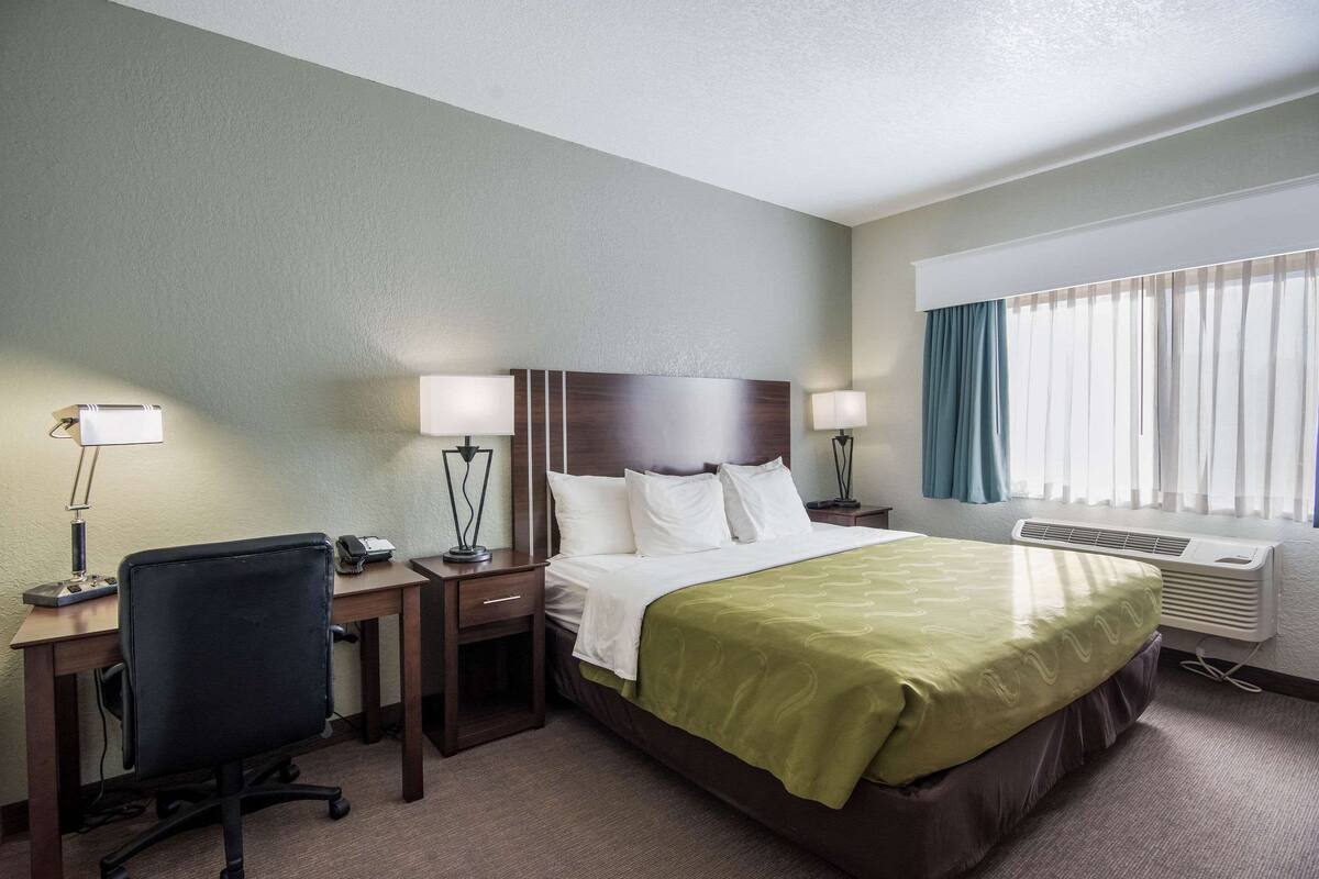 A hotel room with a bed, two bedside tables with lamps, a desk with a chair, and a window with curtains.