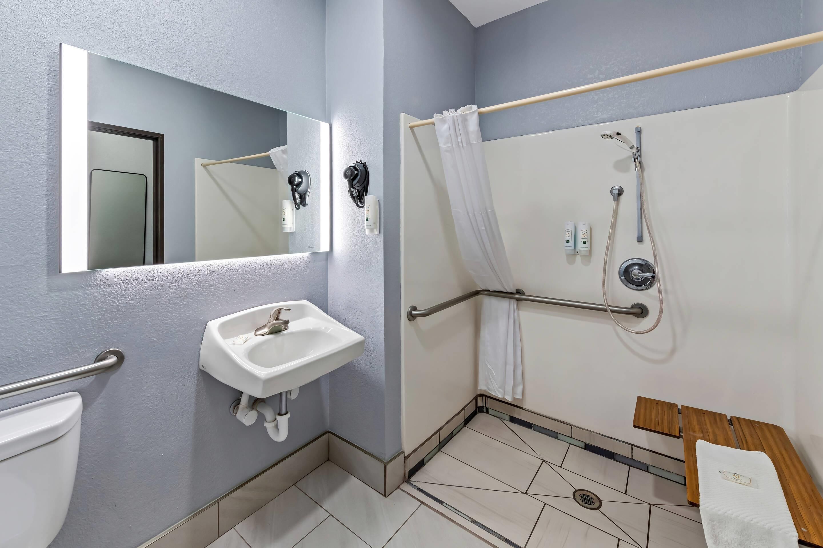 A bathroom with a sink, mirror, shower with grab bars, and a wooden bench.